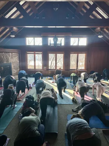 Minnesota Arboretum Fireplace Room with lots of people in dragon pose. Matthew is sitting in the front of the room playing harmonium with 3 big windows behind him.