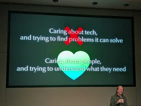 Photo of a presentation slide contrasting “Caring about tech and trying to find problems it can solve” (over a red X) with “Caring about people and trying to understand what they need” (over a green heart), with a speaker holding a microphone at bottom right.