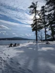 footprints in the snow toward chairs by a lake framed by tall pine trees