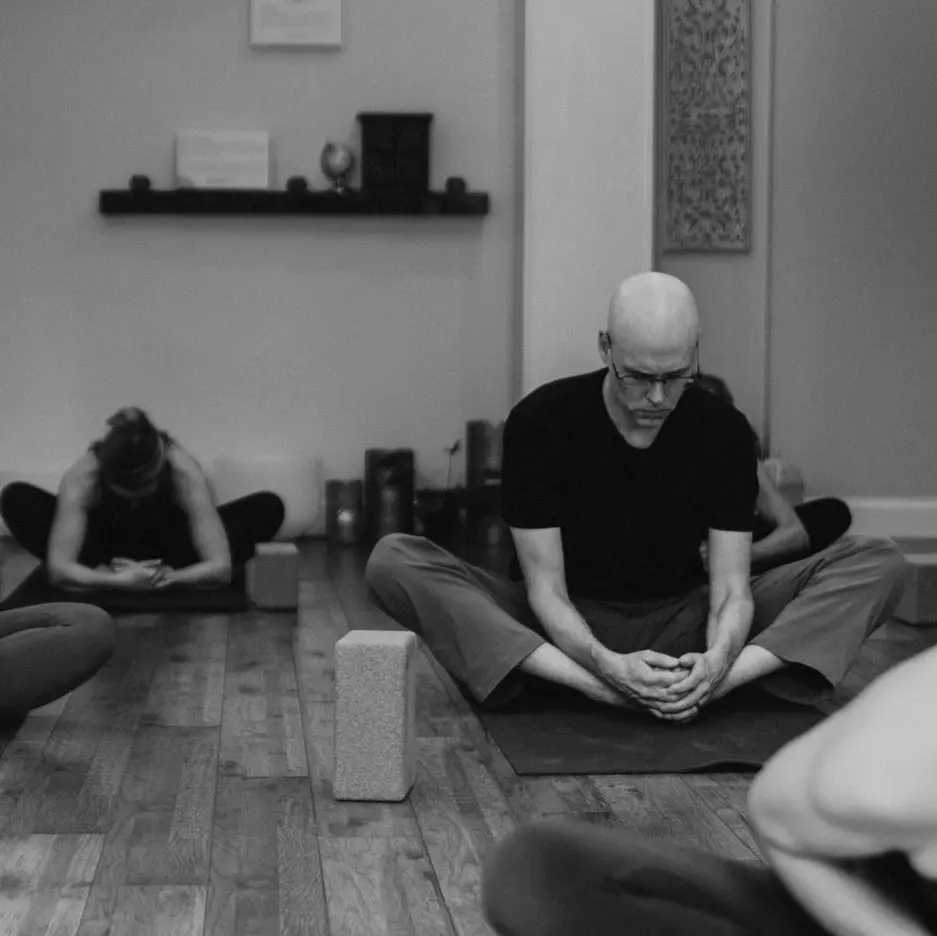 A black and white photo of Matthew sitting in badha konasana in a yoga class