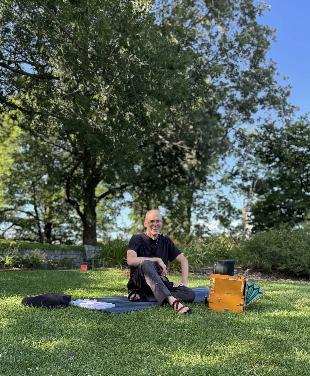 matthing sitting on a yoga mat next to his harmonium in the grass near trees
