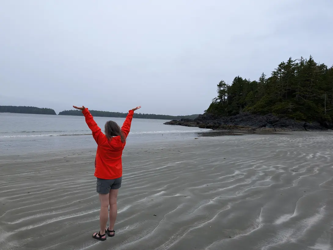 A person in a red jacket stands on a wet sandy beach with arms raised toward an overcast sky, facing the ocean