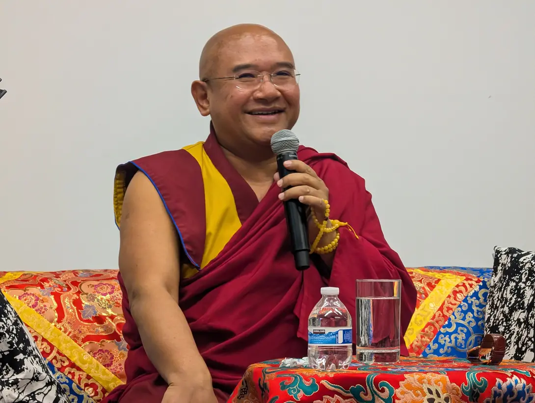 A smiling, shaved-headed Tibetan Buddhist monk wearing traditional maroon and yellow robes. He is wearing glasses and holding a black microphone in his right hand, around which a strand of yellow prayer beads (mala) is wrapped. He is seated on a couch with colorful, ornate, patterned fabric. In front of him, on a small table draped in a matching patterned cloth, sits a clear plastic water bottle and a glass of water. The background is a plain white wall.